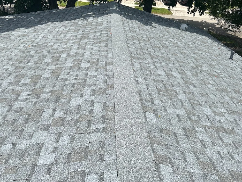 A close-up view of a gray residential roofing shingle roof with a ridge running through the center, surrounded by trees and some Miami sunlight casting shadows on the roof surface.