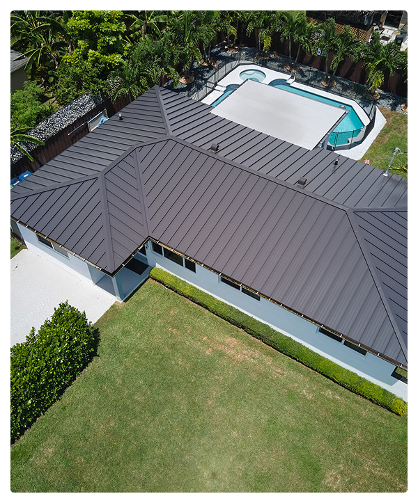 Aerial view of a modern Miami house with a dark metal roofing, manicured lawn, nearby hedge, and a backyard pool with a cover, surrounded by palm trees and fencing—perfectly finished by affordable contractors.