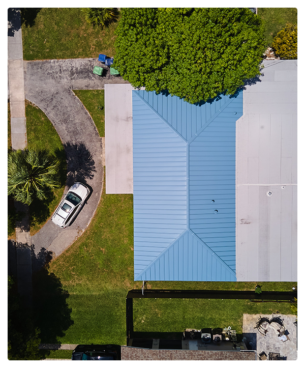 Aerial view of a Miami house with a blue metal roof, green lawn, large tree, and a white car parked on a curved driveway. Two trash bins sit at the edge, highlighting work done by affordable contractors specializing in roofing.
