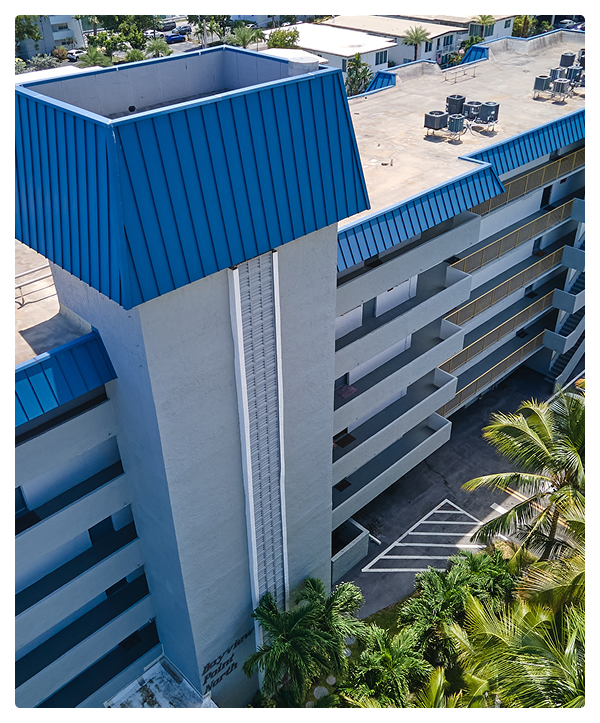 A multi-story building in Miami with blue residential roofing accents, exterior corridors, and an elevator shaft. Palm trees are visible in the foreground, and the rooftop features HVAC units and equipment.