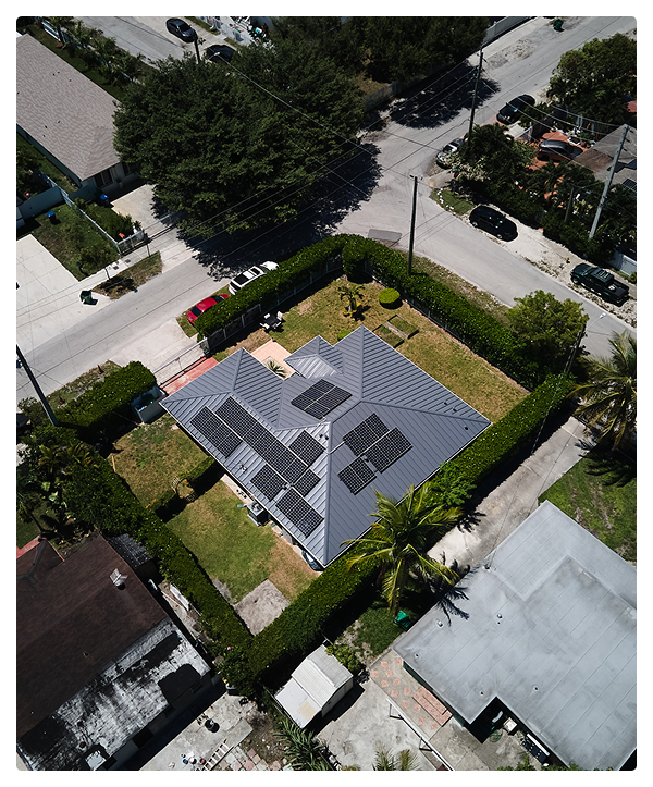 Aerial view of a house with a gray metal roof and several solar panels, surrounded by a green lawn and trees in a Miami neighborhood. Nearby streets, parked cars, and expert affordable contractors add to the vibrant residential scene.