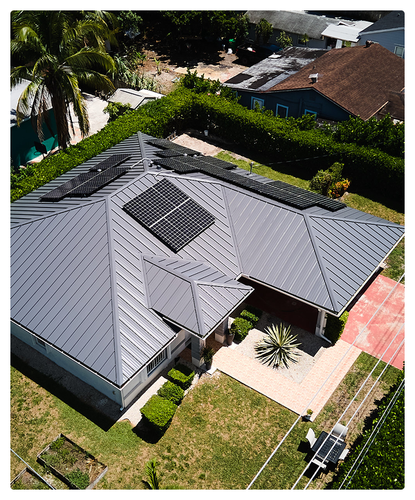 Aerial view of a house with a dark gray metal residential roofing system featuring multiple solar panels, surrounded by a green yard, bushes, and nearby houses.