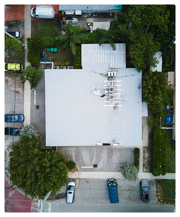 Aerial view of a building with a white commercial roofing surrounded by trees and parked cars. The building, ideal for affordable contractors, is bordered by streets on three sides, with various vehicles visible along the roads and in driveways.