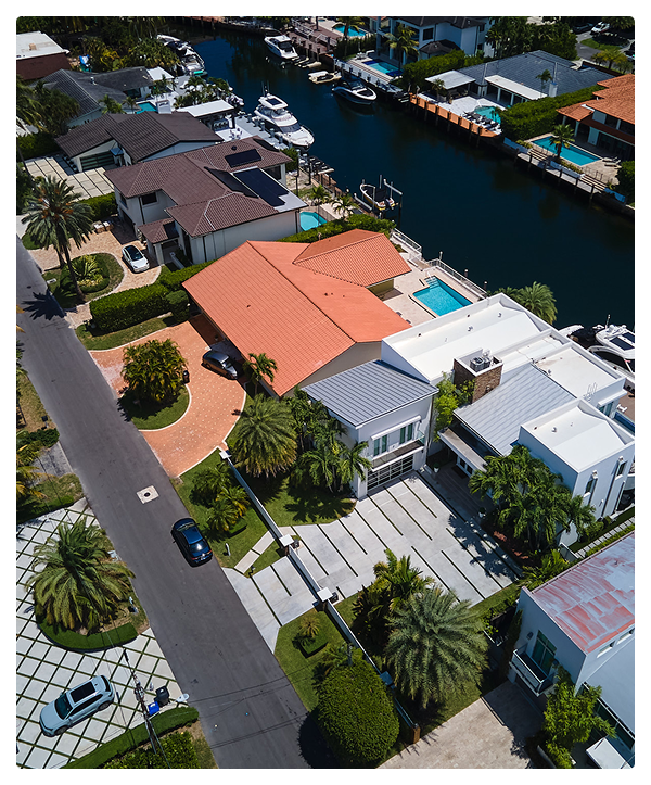 Aerial view of a suburban neighborhood with modern houses featuring quality roofing, palm trees, a canal with boats, swimming pools, and parked cars along the street on a sunny day.