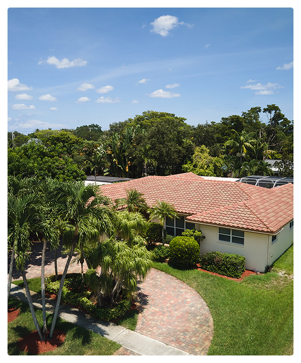 Aerial view of a single-story house with a red-tiled roof, surrounded by palm trees, green lawn, and landscaped bushes under a clear blue Miami sky—quality residential roofing by affordable contractors.