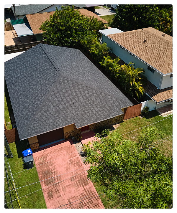Aerial view of a single-story house with a dark gray roofing, red brick driveway, green lawn, and neighboring homes surrounded by trees and shrubs.
