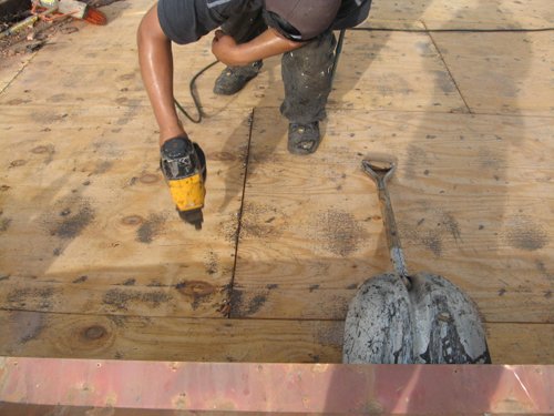 A person using a yellow power drill on a plywood floor at a Miami construction site, with a mud-covered shovel nearby. Only the person’s torso, arms, and legs are visible—perfect for commercial roofing or residential roofing projects.