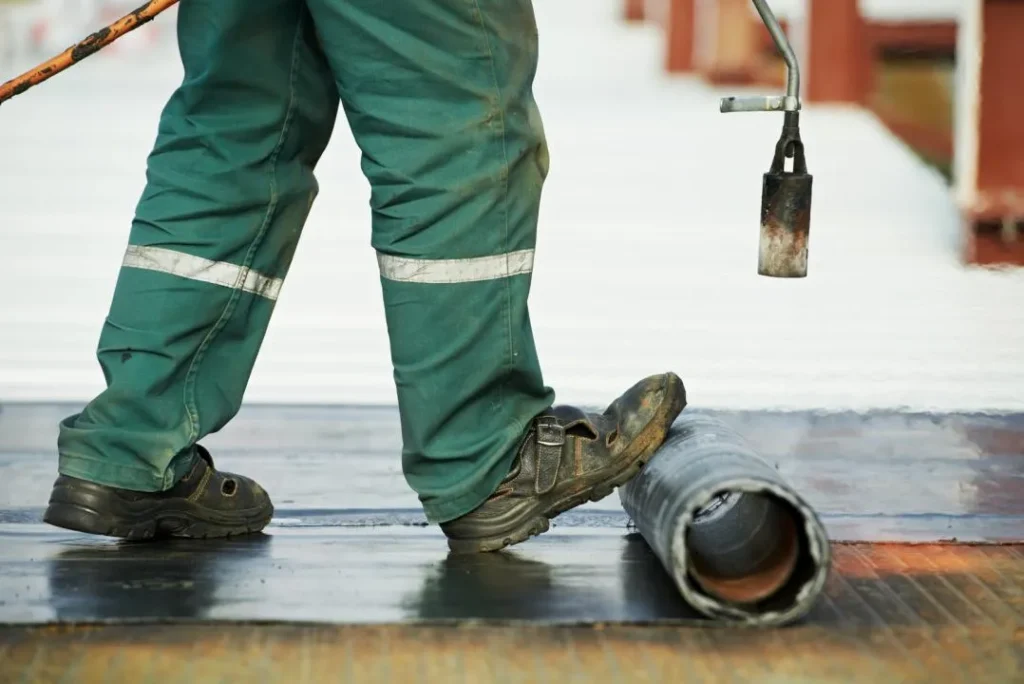A worker in green pants and black boots uses a torch to apply roofing material, unrolling a black sheet onto a flat surface—showcasing quality residential roofing by Affordable Contractors in Miami.