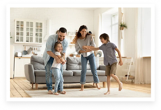 A happy family of four—two adults and two children—laugh and play together in a bright, modern Miami living room with a gray sofa and light wooden flooring. Sunlight streams in from large windows, highlighting their joy.
