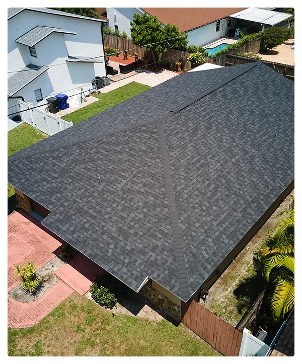 Aerial view of a house with a newly installed dark gray shingle roof by affordable contractors, surrounded by a yard with trees, a brick walkway, and nearby fences and neighboring homes.