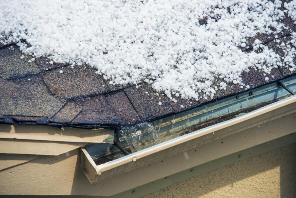 Hailstones cover a shingled roof as water flows into a rain gutter during a hailstorm, highlighting the importance of quality residential roofing in Miami.
