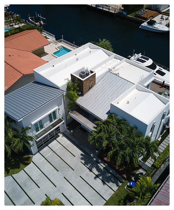 Aerial view of a modern white house with flat roofs by Affordable Contractors, featuring a driveway, palm trees, backyard pool, and canal dock with boats—nestled among other waterfront properties.