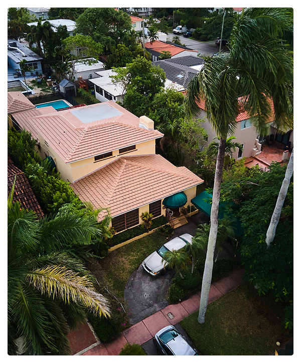Aerial view of a suburban Miami house with a terracotta roof, a curved driveway with two parked cars, tall palm trees, and neighboring houses surrounded by lush greenery—all completed by affordable contractors specializing in roofing.